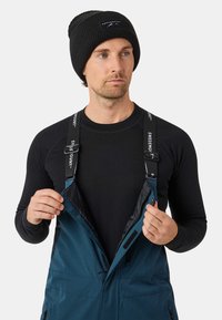Blauwe waterdichte overalls met verstelbare zwarte banden, een zwarte trui eronder en een zwarte beanie. Voorzien van rits- en klittenbandsluitingen.