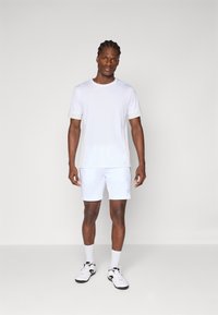 Λευκό μπλουζάκι με κοντό μανίκι και στρογγυλή λαιμόκοψη, συνδυασμένο με λευκά σορτς. Το ύφασμα φαίνεται λείο, σχεδιασμένο για αθλητική χρήση.