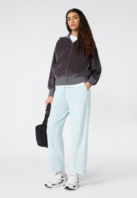 Veste zippée en velours gris, pantalon léger bleu clair à coupe ample, sac bandoulière noir et chaussures de sport blanches avec des accents noirs.
