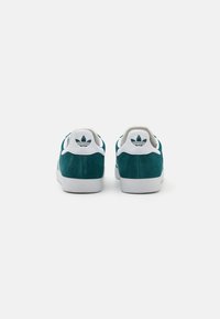 Green suede sneakers featuring a white rubber sole, white accents, and a logo on the heel. Simplistic design with a smooth texture.