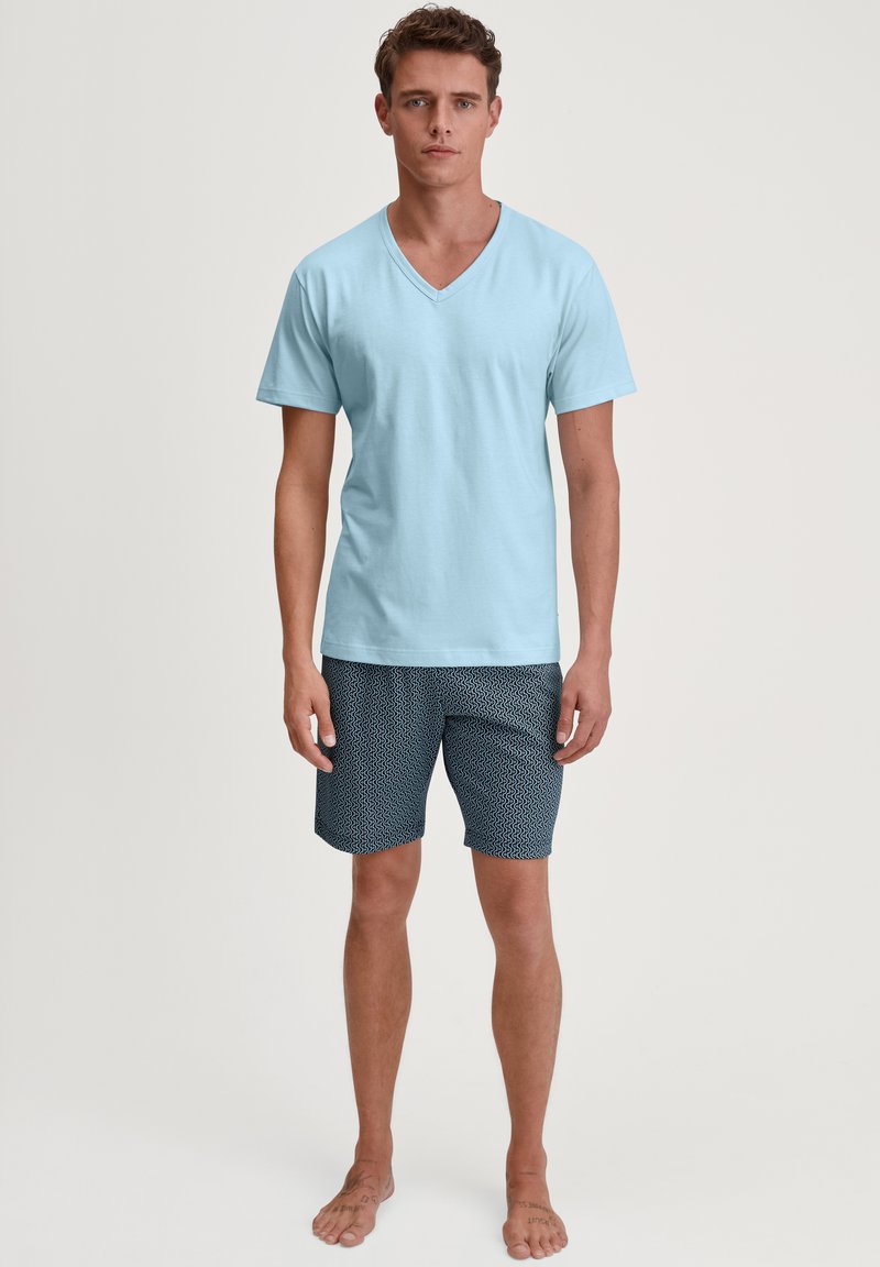 Lichtblauwe v-hals t-shirt gecombineerd met marineblauwe shorts met een wit golfpatroon. Model staat blootsvoets en toont de outfit duidelijk.