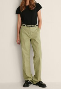 Světlé zelené kalhoty s širokými nohavicemi a plochým pasem, s bočními kapsami a černým pásem. V kombinaci s těsně přiléhajícím černým tričkem a masivními černými botami.