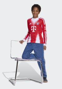 Rood en wit voetbalshirt met lange mouwen, voorzien van een gradient ontwerp, logo en strepen, gecombineerd met blauwe jeans. Duidelijke acrylstoel.