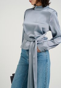 Lys blå satinskjorte med høj hals og lange ærmer, der har et bånd i taljen. Kombinationen er med lys blå denim jeans.