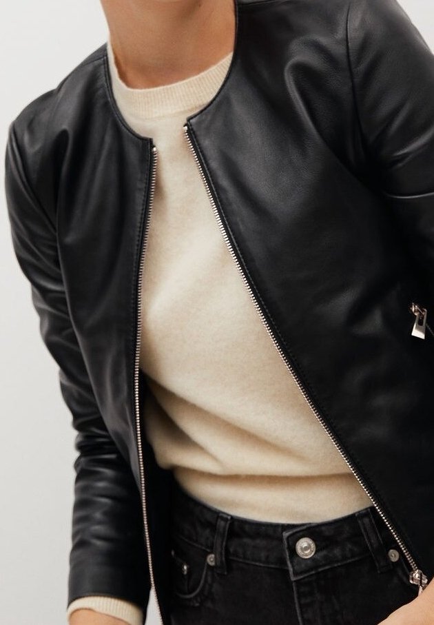 Zwarte leren bomberjack met een ritssluiting, ronde halslijn en aansluitende mouwen, gedragen over een crèmekleurige trui. Gelaagde structuur en een soepele afwerking.