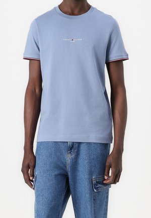 Homme portant un t-shirt bleu clair Tommy Hilfiger avec logo et un pantalon cargo en denim bleu moyen sur fond blanc.