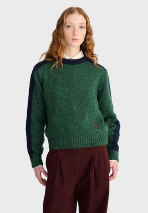Jeune personne aux longs cheveux roux porte un pull en laine vert avec des rayures bleu marine sur les manches et un pantalon rouge foncé, debout devant un fond clair.