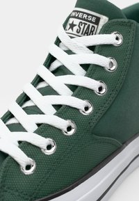 Sapatilhas verdes de lona com atacadores brancos, ilhós prateados e um patch de logo preto e branco. Apresenta uma superfície texturizada e sola de borracha.