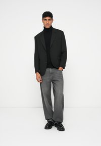 Blazer preto com lapela de corte, usado sobre um suéter preto de gola alta. Combinado com jeans cinza largos e calçado preto. Textura suave, design ajustado.
