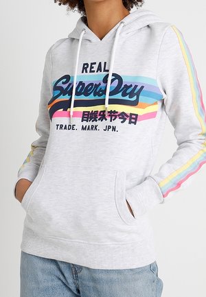Osoba nosząca jasnoszarą bluzę z kapturem Superdry z tęczowymi paskami na rękawach i kolorowym napisem logo na przodzie, ręce w przedniej kieszeni.
