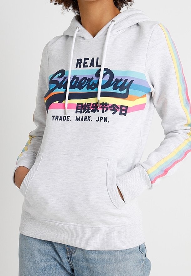 Personne portant un sweat à capuche Superdry gris clair avec des bandes arc-en-ciel sur les manches et un texte de logo coloré à l'avant, mains dans la poche avant.