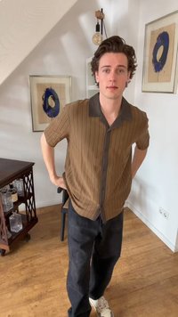 Νέος άνδρας με καφέ ριγέ πουκάμισο και σκούρο παντελόνι στέκεται μέσα σε εσωτερικό χώρο κοντά σε κορνιζαρισμένη μπλε κυκλική τέχνη και ξύλινο μπαρ με ποτήρια.