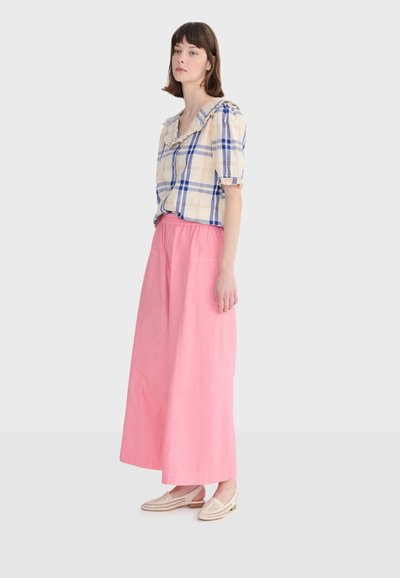 Femme portant un chemisier à carreaux beige avec un col volanté, un pantalon large taille haute rose, et des chaussures beige tissées à enfiler, debout de profil.