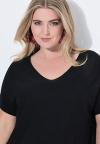 Černý top s výstřihem do V a krátkými rukávy, vyrobený z texturované látky. Modelka má na sobě červené květinové náušnice a má jemné, vlnité vlasy. Neutrální pozadí.