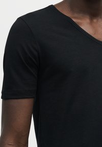 Gros plan d'une personne portant un t-shirt noir à manches courtes avec un col en V, montrant le haut du torse et le bras.