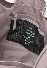 Interno di una borsa a tracolla in tessuto rosa con una tasca con zip e una sezione in rete, con un'etichetta nera che mostra un testo umoristico in carattere bianco.