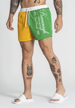 Mężczyzna ubrany w żółto-zielone spodenki kąpielowe z białym sznurkiem i napisem, białe klapki, widoczne liczne tatuaże na ramionach i nogach.