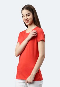 T-shirt rosso a maniche corte in un tessuto morbido, con scollo rotondo e una targhetta laterale, indossato con pantaloni bianchi.