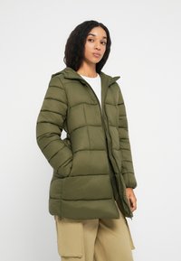 Mujer vistiendo una chaqueta acolchada verde aceituna con capucha, combinada con pantalones cargo beige y una camisa blanca, de pie contra un fondo blanco.