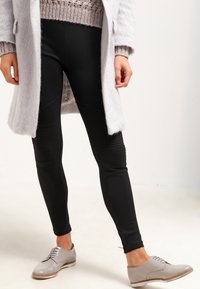 Mustat tiukat legginsit, joissa on tekstuuriset polvisyvennykset, yhdistettynä vaaleanharmaaseen ylisuurikokoiseen takkiin ja harmaisiin nauhakenkiin. Pehmeä neulepusero alla.