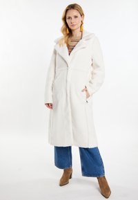Lunga giacca bianca realizzata in materiale soffice, con cappuccio, tasche frontali e design senza cuciture; indossata sopra jeans a gamba larga blu e stivali marroni.