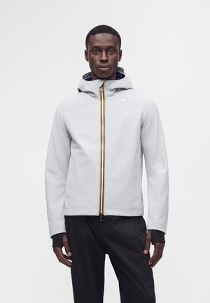 Homme portant une veste à capuche gris clair avec une fermeture éclair jaune et noire, associée à un pantalon noir, debout devant un fond blanc uni.
