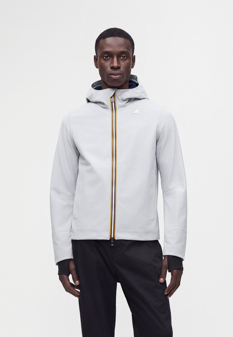 Homme portant une veste à capuche gris clair avec une fermeture éclair jaune et noire, associée à un pantalon noir, debout devant un fond blanc uni.