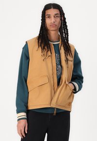 Gilet di jeans color nocciola con due tasche frontali, design aperto, indossato sopra un felpa verde con un logo circolare e polsini a righe.