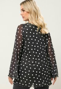 Blusa nera trasparente con pois bianchi, maniche a campana lunghe e orlo scalloppato, dotata di scollatura rotonda e vestibilità morbida.