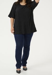 Femme portant un haut noir texturé à manches courtes, un jean bleu foncé et des chaussures noires à talons pointus avec détail de boucle en argent, se tenant sur fond blanc.