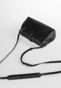 Borsa a tracolla in pelle nera con una forma arrotondata, dotata di chiusura a pattina, tracolla regolabile e un leggero branding sul lato.
