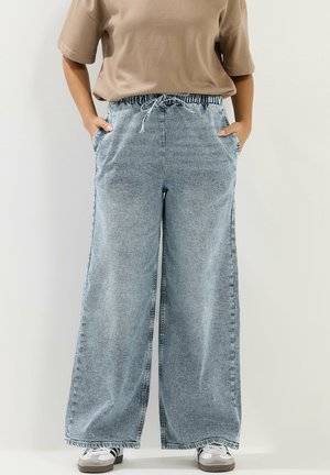 Person iført lyseblå vidde cowboybukser med snøre i taljen, beige kortærmet top, hænderne i lommerne og hvide sneakers.