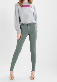 Sudadera gris con texto rojo, combinada con jeans ajustados verdes. Modelo vistiendo tacones altos dorados brillantes. Diseño simple, forma ajustada.