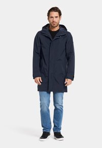 Marineblaue wasserdichte Jacke mit Kapuze, Knopfverschluss und Seitentaschen; kombiniert mit hellblauen Jeans und schwarzen Sneakern. Glatte Textur.