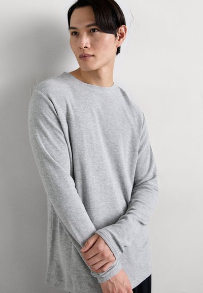Hellgraues Langarm-T-Shirt aus weichem Stoff, mit rundem Ausschnitt und gerippten Bündchen, getragen mit gefalteten Händen vor dem Körper.