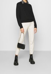 Svart tröja med långa ärmar över en krag, mönstrad skjorta, i kombination med krämfärgade byxor och kraftiga svarta ankelstövlar. Håller en svart handväska.