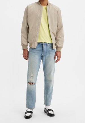 Mand iført beige bomberjakke, gul t-shirt, lyseblå revnede jeans, hvide sokker og sort-hvide loafers, der står mod en hvid baggrund.