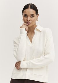 Hvid strikket pullover med krave, v-hals og ribbede manchetter. Den har en løs pasform og en lomme foran, hvilket giver en blød tekstur.