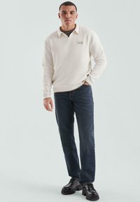 Jonge man die staat in een witte lange mouwen polo, donkere jeans en zwarte stevige schoenen tegen een effen lichte achtergrond.