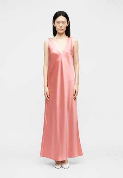 Γυναίκα όρθια, ντυμένη με ροζ σατέν φόρεμα χωρίς μανίκια, μακρύ μέχρι το πάτωμα, με βαθύ V λαιμόκοψη και λευκές μυτερές γόβες, απλό φωτεινό φόντο.