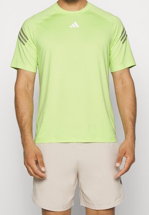 Mand iført en lysegrøn atletisk t-shirt med korte ærmer og grå striber på ærmerne samt beige shorts, står med armene afslappet ned langs siden.