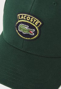 Grön bomullscap med strukturerad tyg, böjd skärm och en cirkulär Lacoste-logotyp-etikett med en broderad krokodil-design.
