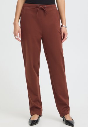 Femme portant un pantalon marron à jambes droites avec une taille à cordon et des chaussures noires à petits talons, debout sur un fond clair.