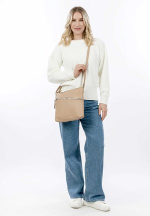 Tan synthetic crossbody bag with a zippered front pocket and a long adjustable strap, paired with light blue wide-leg jeans and a white sweater.