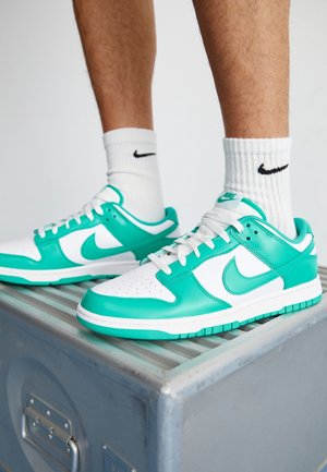 Personne portant des baskets Nike blanches et turquoise avec des chaussettes blanches Nike, debout sur une surface métallique avec des poignées.