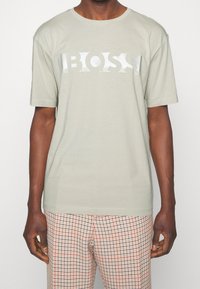 Muž má na sobě světle šedé tričko s textem "BOSS" a béžové kostkované kalhoty s červenými a černými pruhy.