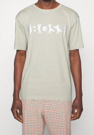 Homme portant un T-shirt gris clair avec le texte "BOSS" et un pantalon à carreaux beige avec des lignes rouges et noires.