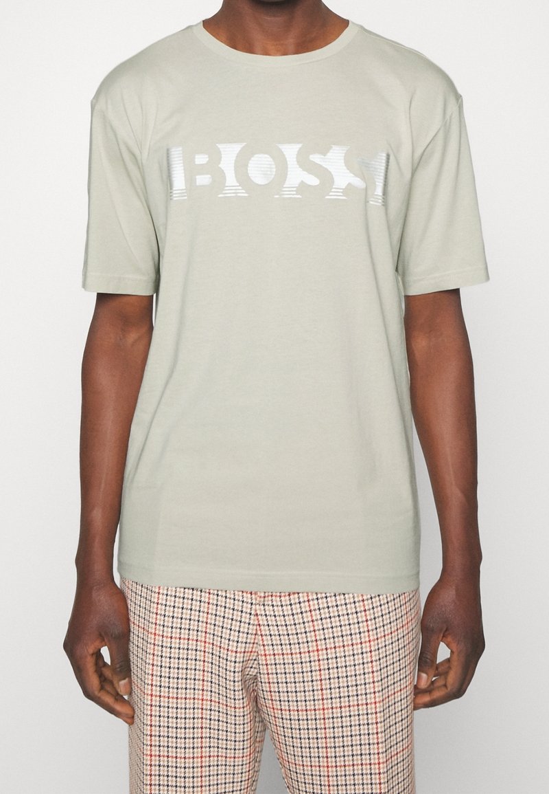 Muž má na sobě světle šedé tričko s textem "BOSS" a béžové kostkované kalhoty s červenými a černými pruhy.