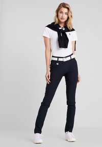 Chemise polo blanche avec un pull noir noué autour des épaules, pantalon bleu marine foncé, ceinture blanche et baskets blanches. Conception simple, style décontracté.