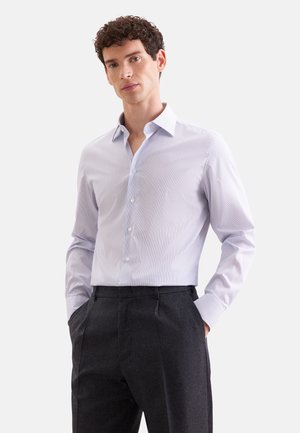 Homme aux cheveux bouclés portant une chemise bleu clair boutonnée et un pantalon gris foncé, les mains dans les poches, sur un fond uni.